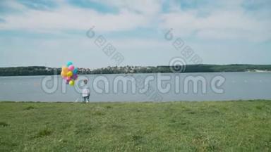 快乐的小男孩站在湖岸上，手里拿着五颜六色的气球。 4K